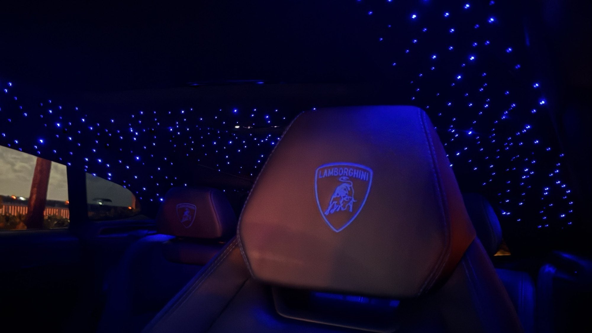 Inside the Urus at night: a starlight headliner of hundreds of small blue lights above black leather seats, with the Lamborghini shield glowing on a headrest