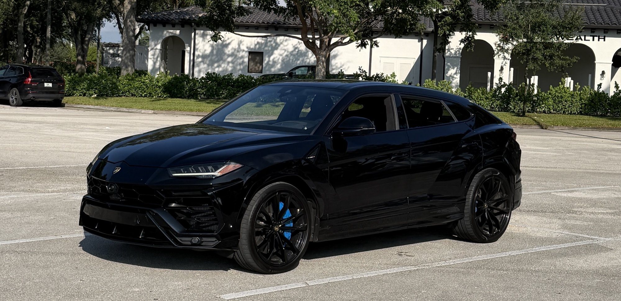 Black Lamborghini Urus seen from the front three-quarter angle, with black wheels and blue brake calipers, parked in an open lot in front of a white building and trees