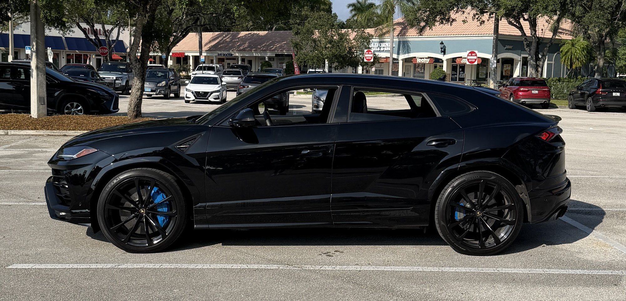 Side profile of a black Lamborghini Urus with tinted windows, large black wheels and blue brake calipers, parked in a shopping plaza lot on a sunny day